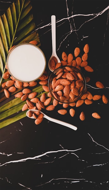 A glass of almond milk is placed on a dark marble surface surrounded by scattered almonds and a palm leaf. A clear bowl filled with almonds is next to the glass, with two white spoons resting nearby, one filled with almonds.