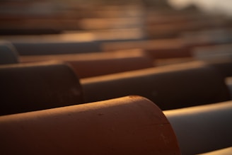 Rows of large industrial pipes are arranged in parallel, with a focus on warm and soft lighting that creates a gentle gradient across the surfaces. The pipes display smooth, rounded edges with uniformly distributed highlights and shadows, suggesting a late afternoon or early evening sun.