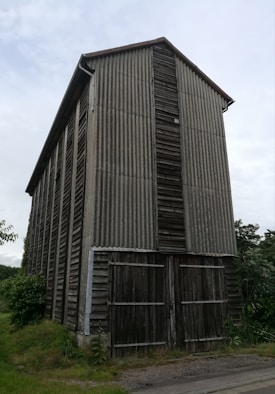 A tall, rectangular wooden barn with vertical corrugated metal siding and a pitched roof. The barn appears weathered and aged, with visible wooden doors at the base. It is surrounded by greenery, including grass and bushes, under an overcast sky.
