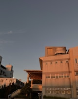 A large building with a modern architectural design is illuminated by the warm hues of the sunset. The structure features multiple windows, a prominent cylindrical structure on the roof, and a large overhang supported by columns. The surrounding area includes trees and shrubs, adding a touch of greenery.