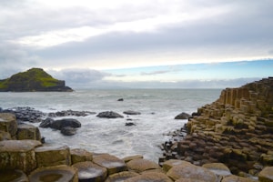 A scenic coastal landscape featuring the Giant's Causeway, characterized by its distinctive hexagonal basalt columns. These natural formations appear to extend into the sea, with waves gently crashing against them. The sky is partly cloudy, creating a serene and picturesque atmosphere.
