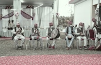 A group of musicians dressed in traditional attire, sitting on chairs and playing various instruments such as drums and horns. The setting appears to be a decorated outdoor venue, with floral arrangements and draped fabric in the background. Some of the musicians wear face masks.