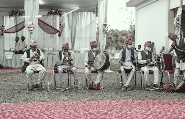 A group of musicians dressed in traditional attire, sitting on chairs and playing various instruments such as drums and horns. The setting appears to be a decorated outdoor venue, with floral arrangements and draped fabric in the background. Some of the musicians wear face masks.