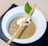 A bowl of creamy mushroom soup garnished with a swirl of cream, a slice of mushroom, a basil leaf, and a long breadstick resting across the top. The soup is served in a white bowl on a black slate surface, creating a contrast with the beige background.