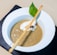 A bowl of creamy mushroom soup garnished with a swirl of cream, a slice of mushroom, a basil leaf, and a long breadstick resting across the top. The soup is served in a white bowl on a black slate surface, creating a contrast with the beige background.