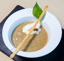 A bowl of creamy mushroom soup garnished with a swirl of cream, a slice of mushroom, a basil leaf, and a long breadstick resting across the top. The soup is served in a white bowl on a black slate surface, creating a contrast with the beige background.