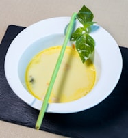 A white bowl containing creamy yellow soup is set on a dark slate surface. There is a lemongrass stalk and fresh green basil leaves garnishing the soup.