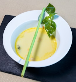 A white bowl containing creamy yellow soup is set on a dark slate surface. There is a lemongrass stalk and fresh green basil leaves garnishing the soup.