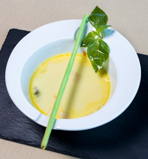 A white bowl containing creamy yellow soup is set on a dark slate surface. There is a lemongrass stalk and fresh green basil leaves garnishing the soup.
