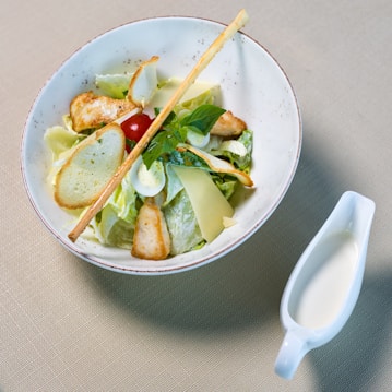 A bowl of salad featuring lettuce, cherry tomatoes, croutons, slices of grilled chicken, hard-boiled egg slices, cheese, and a grissini breadstick. A small jug of creamy dressing is placed beside the bowl on a textured beige surface.
