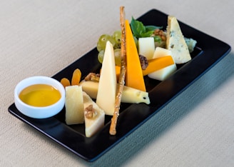 An elegantly arranged cheese platter on a black rectangular plate, featuring a variety of cheese slices, including cheddar, blue cheese, and brie. Accompanying the cheeses are walnuts, dried apricots, green grapes, and a crispy breadstick. A small white bowl filled with honey or olive oil sits next to the cheeses. The background is a simple beige fabric.