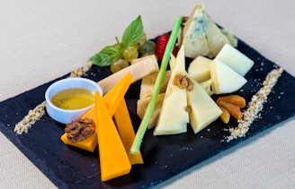 Assorted cheese slices arranged on a black slate board, complemented by nuts, dried fruits, green grapes, and fresh basil. A small dish containing honey or oil is also present. The cheeses vary in color and type, with some garnished with walnut halves.