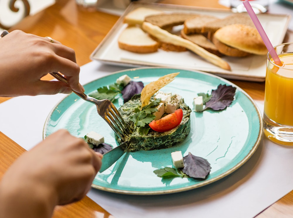 personne tenant une fourchette et un couteau coupant des légumes sur une plaque en céramique blanche