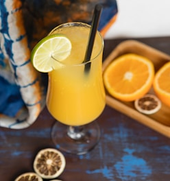 A refreshing yellow-orange drink in a tall glass with a black straw, garnished with a fresh lime slice. There are halved oranges placed in a wooden tray and additional slices of dried citrus fruit scattered on the blue and brown table. A colorful fabric with shades of blue and orange is draped in the background.