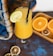 A refreshing yellow-orange drink in a tall glass with a black straw, garnished with a fresh lime slice. There are halved oranges placed in a wooden tray and additional slices of dried citrus fruit scattered on the blue and brown table. A colorful fabric with shades of blue and orange is draped in the background.