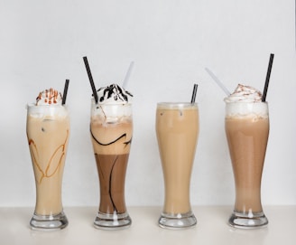 Four tall glasses filled with creamy iced beverages, each topped with whipped cream and sauces. The first and second drinks have caramel and chocolate drizzles respectively, while the third and fourth appear to be simple iced coffees. Straws are placed in each glass.