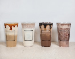 Four plastic cups are filled with various iced beverages, each topped with different types of foam or drizzles. The drinks have varying shades and textures, suggesting a mix of coffee or tea-based drinks, possibly with milk or cream. They are neatly arranged in a row on a light-colored surface with minimalistic labels.