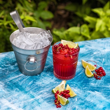 A metal bucket filled with ice cubes sits on a textured blue surface next to a vibrant red drink garnished with lime wedges and clusters of red currants. Additional lime wedges and red currants are scattered around, with a lush green foliage backdrop.