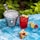 A metal bucket filled with ice cubes sits on a textured blue surface next to a vibrant red drink garnished with lime wedges and clusters of red currants. Additional lime wedges and red currants are scattered around, with a lush green foliage backdrop.