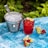 A metal bucket filled with ice cubes sits on a textured blue surface next to a vibrant red drink garnished with lime wedges and clusters of red currants. Additional lime wedges and red currants are scattered around, with a lush green foliage backdrop.