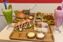 A variety of dishes are laid out on wooden platters, featuring a burger with lettuce and tomato, a hot dog drizzled with mayonnaise, a wrap with mixed fillings, and an assortment of dips and side salads. There are two vibrant smoothies, one green with a kiwi slice and another pink with a strawberry on top. Fried falafel balls and fresh sliced vegetables are also present, creating a colorful and appetizing display.
