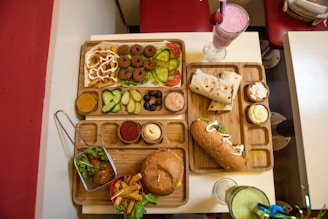 An assortment of various foods is served on wooden trays on a table. On one tray, there are falafel balls, slices of cucumber, pickle, olives, and sauces. Another tray includes a wrap and a sub sandwich, accompanied by small servings of different sauces and a milkshake with a pink hue topped with a strawberry. A basket with fried appetizers and a burger with fries is arranged on a separate tray. Drinks in clear glasses are also present, with pink and green beverages visible.