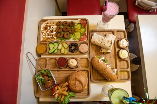 An assortment of various foods is served on wooden trays on a table. On one tray, there are falafel balls, slices of cucumber, pickle, olives, and sauces. Another tray includes a wrap and a sub sandwich, accompanied by small servings of different sauces and a milkshake with a pink hue topped with a strawberry. A basket with fried appetizers and a burger with fries is arranged on a separate tray. Drinks in clear glasses are also present, with pink and green beverages visible.