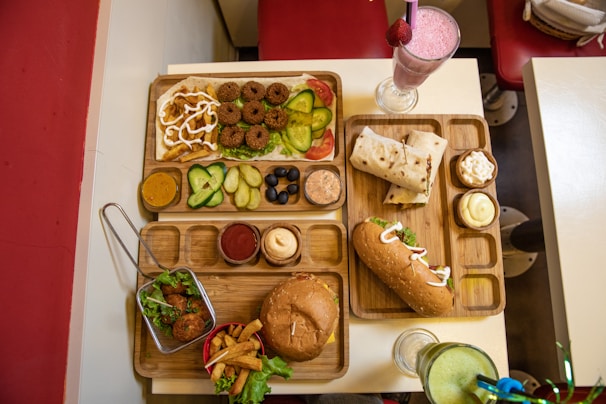 An assortment of various foods is served on wooden trays on a table. On one tray, there are falafel balls, slices of cucumber, pickle, olives, and sauces. Another tray includes a wrap and a sub sandwich, accompanied by small servings of different sauces and a milkshake with a pink hue topped with a strawberry. A basket with fried appetizers and a burger with fries is arranged on a separate tray. Drinks in clear glasses are also present, with pink and green beverages visible.