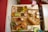An assortment of various foods is served on wooden trays on a table. On one tray, there are falafel balls, slices of cucumber, pickle, olives, and sauces. Another tray includes a wrap and a sub sandwich, accompanied by small servings of different sauces and a milkshake with a pink hue topped with a strawberry. A basket with fried appetizers and a burger with fries is arranged on a separate tray. Drinks in clear glasses are also present, with pink and green beverages visible.