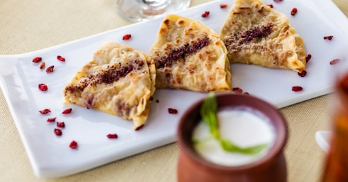 Three triangular pastries with a flaky, golden-brown crust are arranged neatly on a white rectangular plate. The pastries have a visible filling and are garnished with red flakes. Scattered around the pastries are small red berries, adding a pop of color. In front of the plate, there is a small clay cup containing a white liquid with a green garnish.