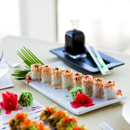 A neatly arranged plate of sushi rolls garnished with sauces, green herbs, and slices of ginger. A small bottle containing a dark liquid, likely soy sauce, sits in the background alongside a small tray with chopsticks. The table is set on a light-colored tablecloth, and additional sushi pieces with colorful toppings are partially visible in the foreground.