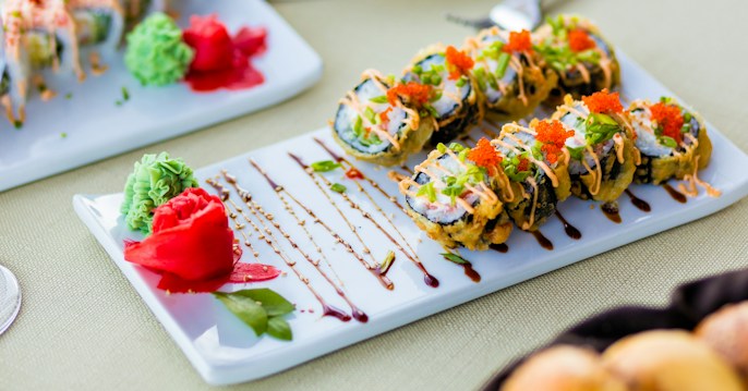 A platter of sushi rolls garnished with vibrant orange fish roe, creamy drizzle, and finely chopped green herbs. Accompaniments include vividly colored wasabi and bright red pickled ginger. The elegant presentation is enhanced with artistically arranged sauce lines on the plate.
