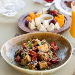 A close-up of a delicious meal featuring a bowl of sautéed tofu with cherry tomatoes and garnished with sesame seeds and fresh herbs. In the background, there is a platter with a variety of cheeses and fruits, while a glass of orange juice is placed on the right, enhancing the dining ambiance.