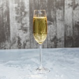 A tall, elegant champagne glass filled with sparkling golden liquid is placed on a light, textured surface. The background features a blurred, gray textured wall, enhancing the contrast with the clear glass and the bubbles rising inside.