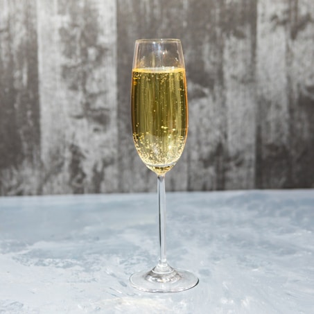 A tall, elegant champagne glass filled with sparkling golden liquid is placed on a light, textured surface. The background features a blurred, gray textured wall, enhancing the contrast with the clear glass and the bubbles rising inside.