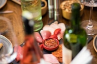 An elegant spread of cured meats and sliced cheese is laid out on a wooden table. A small red bowl contains a relish or dip. The table also features bottles of wine and possibly sparkling water, along with polished cutlery and plates, suggesting a refined dining setup. The warm colors and soft focus create an inviting and cozy atmosphere.