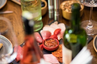 An elegant spread of cured meats and sliced cheese is laid out on a wooden table. A small red bowl contains a relish or dip. The table also features bottles of wine and possibly sparkling water, along with polished cutlery and plates, suggesting a refined dining setup. The warm colors and soft focus create an inviting and cozy atmosphere.