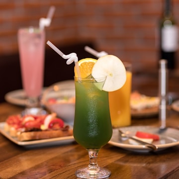 A close-up view of a table with assorted drinks and food. The focal point is a green cocktail in a stemmed glass, garnished with an orange slice and an apple slice, and two white straws. Behind it, a pink drink in a tall glass and an orange drink are visible, along with plates of bruschetta topped with tomatoes.
