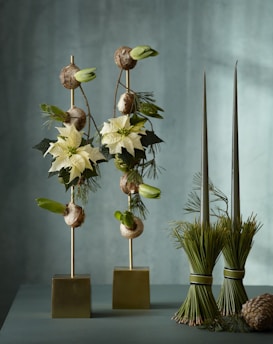 A sophisticated floral arrangement featuring two vertical structures with white poinsettias, greenery, and bulbs, set against a soft green backdrop. Two bundles of tied pine needles stand beside the flowers, adding an organic touch.