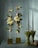 A sophisticated floral arrangement featuring two vertical structures with white poinsettias, greenery, and bulbs, set against a soft green backdrop. Two bundles of tied pine needles stand beside the flowers, adding an organic touch.