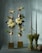 A sophisticated floral arrangement featuring two vertical structures with white poinsettias, greenery, and bulbs, set against a soft green backdrop. Two bundles of tied pine needles stand beside the flowers, adding an organic touch.