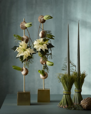 A sophisticated floral arrangement featuring two vertical structures with white poinsettias, greenery, and bulbs, set against a soft green backdrop. Two bundles of tied pine needles stand beside the flowers, adding an organic touch.