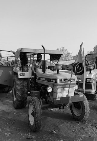 A monochrome photograph featuring a tractor with the brand name Sonalika visible on the front. The tractor is parked outdoors with a flag attached to it. The background includes another similar vehicle and some scattered leaves or debris on the ground. A person is seated on the tractor, dressed in casual outdoor attire.