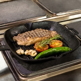 A cooked steak with grill marks is set in a black skillet. Alongside the steak, there are two grilled tomato halves, a green chili pepper, a small bunch of rosemary, and garlic cloves. The skillet is placed on a stovetop.