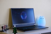 A modern Alienware laptop is open and displaying a glowing alien-themed wallpaper onscreen. To the right of the laptop, there is a cute, glowing cat-shaped lamp with a serene expression. On the left side, small collectible figures are encased in transparent domes on the desk, alongside a small model tree.