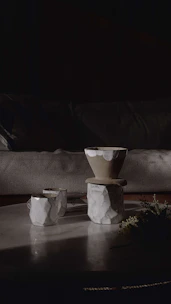 Softly lit ceramic mugs and bowls arranged on a light wood table with natural linen textiles nearby.