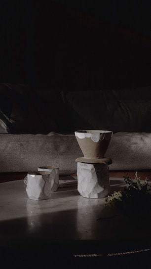 Ceramic coffee cups and bowls with a textured pattern are arranged on a round marble table. Soft lighting casts gentle shadows, creating a cozy and inviting atmosphere. A small plant rests beside the cups.