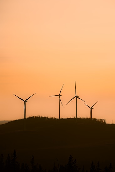 Silhouettes of four wind turbines stand on a hill against a backdrop of a warm, orange sunset. The landscape is minimalistic, with trees at the base of the hill, and the sky has a gradient from orange to a lighter hue.
