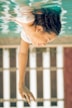 A person with long dark hair appears to be floating or swimming upside down in clear blue water. The image captures a calm and serene moment with the person's eyes closed and one arm extended forward. The background features blurred vertical lines, possibly part of a building or fence, adding to the tranquil atmosphere.