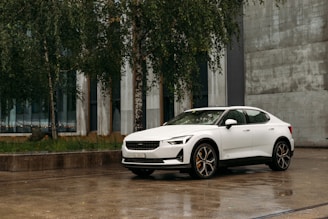 A sleek white electric car is parked on a wet concrete surface surrounded by some trees. The background shows a modern building with large windows and a rough concrete wall. The setting appears to be urban, possibly indicating a recent rain due to the wet ground.